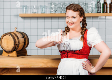 Oktoberfest Kellnerin im traditionellen bayerischen Kleid, "Daumen hoch" Geste und stehen in der Nähe der Theke mit Barrel Bier Stockfoto