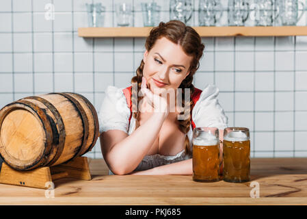 Lächelnd oktoberfest Kellnerin in traditionellen deutschen Kleid stehen an der Theke mit Barrel Bier und zwei Becher helles Bier Stockfoto