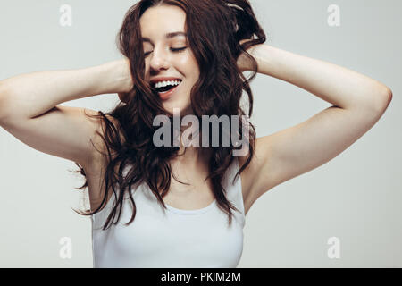 Frau in fröhliche Stimmung lachend mit geschlossenen Augen und die Hände im Haar. Nahaufnahme einer jungen Frau in Casuals auf grauem Hintergrund. Stockfoto