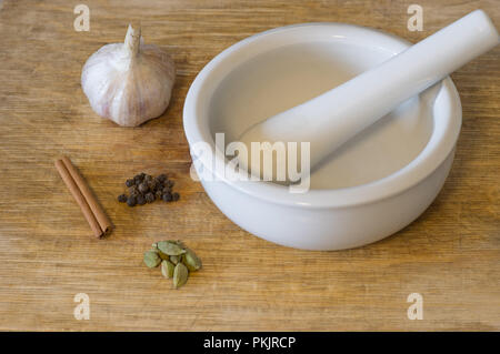 Kardamom, schwarzer Pfeffer, Zimt und Knoblauch liegen in der Nähe von Mörtel auf einem Schneidebrett Stockfoto