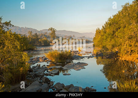 Atemberaubende australische Landschaft mit Felsen verstreut blaue Wasser von Mann Fluss gesäumt von Wäldern am Fuße der Bereiche unter blauem Himmel des frühen Morgens in NSW Stockfoto