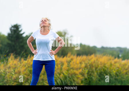 Erwachsene Frau mit Händen auf den Hüften nach oben beim Tragen der blauen Hosen und weißen T-Shirt mit verschwommenen gelb Blumen im Hintergrund Stockfoto