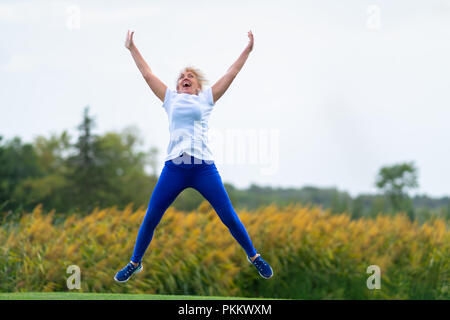 Ältere Frau tragen workout Outfit vor Freude hüpfend mit Armen und Beinen in der Luft vor blurry gelbe Blüten verbreiten Stockfoto