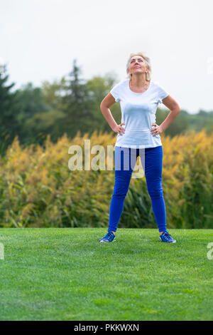 Reife Frau mit blauen Hosen und weißen Hemd mit Zuversicht auf den Hüften vor Blumen Stockfoto