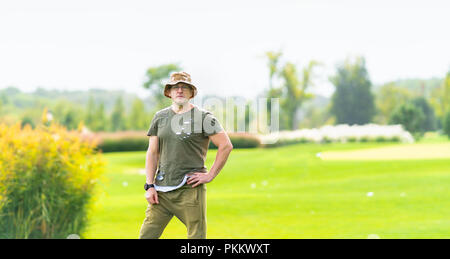 Erwachsener Mann mit dunklen grünen Hemd und braune Hosen stehen in offenen Wiese mit Stückchen Sand im Hintergrund Stockfoto