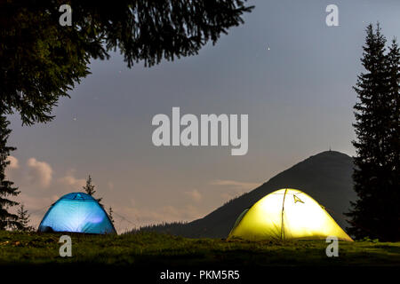 Zwei hell leuchtende, touristische Zelte auf der grünen Wiese Wald Lichtung auf dunklen Berg und dem klaren, blauen Sternenhimmel Kopie Raum Hintergrund. Tourismus, Nacht campin Stockfoto