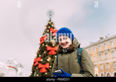 Vilnius, Litauen. Junge schöne Hübsche kaukasische Mädchen Frau gekleidet in grüne Jacke und Blaue Hut Spaß haben und Sie ihr Smartphone auf Hintergrund Chr Stockfoto