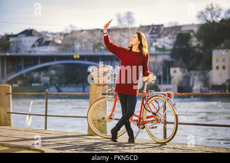 Eine schöne junge Frau mit einem retro rot Fahrrad macht ein Foto von sich in der alten Stadt Europas am Rhein Damm in der Schweizer cit Stockfoto