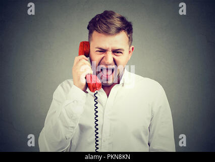 Junger Mann im weißen Hemd mit roten Telefonhörer und Schreien in der Frustration auf grauem Hintergrund. Stockfoto