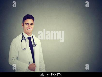 Junge lächelnde männlicher Arzt im weißen Kittel und Stethoskop auf Kamera auf grauem Hintergrund Stockfoto