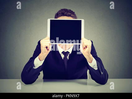 Anonyme Geschäftsmann in Anzug holding Tablet vor Gesicht sitzen auf grauem Hintergrund Stockfoto