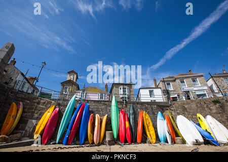 Bunte Kajaks, Kanus und Surfbretter steht aufrecht auf dem sandigen Strand von Mousehole in Cornwall, UK mit dem malerischen Fischerdorf hinter sich. Stockfoto