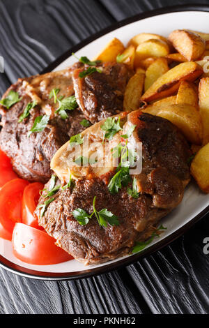 Köstliche Kalbssteak Ossobuco mit Kartoffelscheiben und Tomaten close-up auf einem Teller auf den Tisch. Vertikale Stockfoto