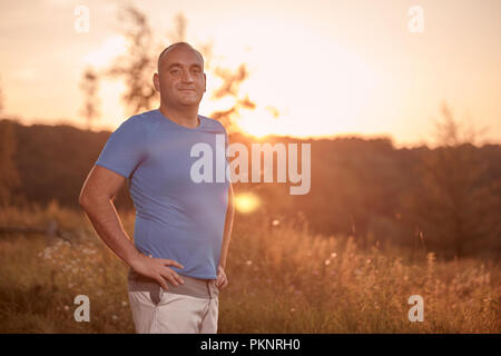 Einem jungen übergewichtigen Mann, 30-35 Jahre, Stolz, ständigen posiert, golden orange Sonnenuntergang, Sonne, im freien Natur Landschaft, ländliche Gegend. Stockfoto