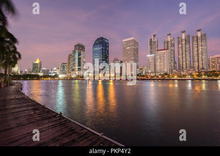 Malerischer Blick auf Holzsteg und See an der Benjakiti (benjakitti) Park und beleuchteten Wolkenkratzer in Bangkok, Thailand, bei Sonnenuntergang. Stockfoto
