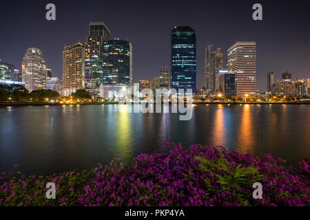 Malerischer Blick auf Blumenbeete und See an der Benjakiti (benjakitti) Park und beleuchteten Wolkenkratzer in Bangkok, Thailand, am Abend. Stockfoto