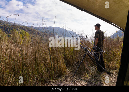 Angeln Abenteuer, Karpfen angeln im Herbst. Fischer mit Camouflage shirt wartet am Ufer eines Sees, versteckt hinter dem Schilf Stockfoto