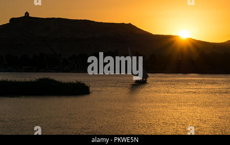 Silhouette des traditionellen Felucca-Segelbootes mit orangefarbenem Sonnenuntergang mit Bergdenkmal Qubbet el Hawa, Nil, Assuan, Ägypten, Afrika Stockfoto