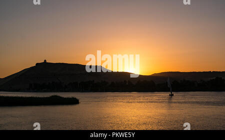 Silhouette der traditionellen Feluke Segelboot mit orange Sonnenuntergang hinter dem Hügel und Hilltop denkmal Qubbet el-Hawa, Nil, Assuan, Ägypten, Afric Stockfoto