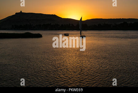 Silhouetten der traditionellen Feluke segeln Boote bei Sonnenuntergang im Nil, Assuan mit hilltop Denkmal Qubbet el-Hawa, Ägypten, Afrika Stockfoto