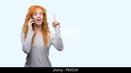 Junge rothaarige Frau sprechen über das Telefon mit einer Idee oder Frage Zeigefinger mit glücklichen Gesicht überrascht, Nummer eins Stockfoto