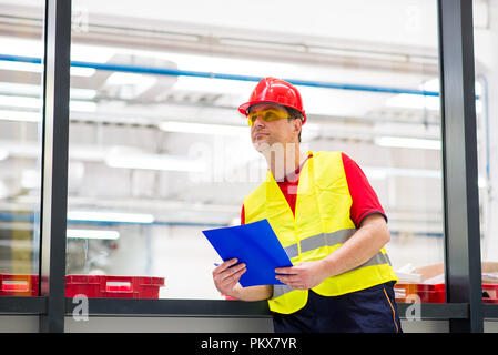 Factory Supervisor in Gelb reflektierende Westen mit roten Helm. Factory Ingenieur holding Blaue Mappe mit Dokumenten Stockfoto