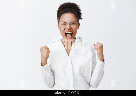 Ja, Mädels, wir haben es geschafft. Portrait von Siegen gut aussehende afrikanische Frau in weißem Hemd und Brille, geballte Fäuste und Kreischen von Erstaunen und Freude, feiern Sieg über graue Wand Stockfoto