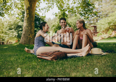 Zwei Frauen im Bikini ihre Getränke Toasten mit einem männlichen Freund in einem Park sitzen. Freunde genießen und entspannen Sie im Park mit Drinks in der Hand. Stockfoto