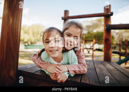 Kleine Mädchen ihre Zwillingsschwester umarmt, während in einem hölzernen Struktur am Spielplatz liegt. Schöne Mädchen genießen im Park spielen. Stockfoto