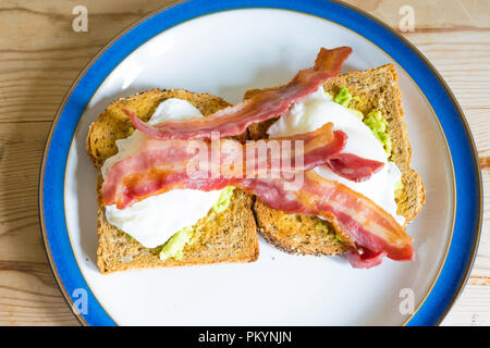 Mittag Sandwich aus zwei pochierte Eier auf Avocado und gerösteten Mehrkornbrot gekrönt mit gegrillten Geräucherter durchwachsener Speck Speckscheiben Stockfoto