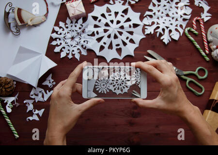 Das Konzept des Neuen Jahres. Eine Frau, die ein Foto für ein Blog. Frauen Hände halten ein Smartphone, Foto Schneeflocken aus Papier, Geschenke, Schere auf einer Stockfoto