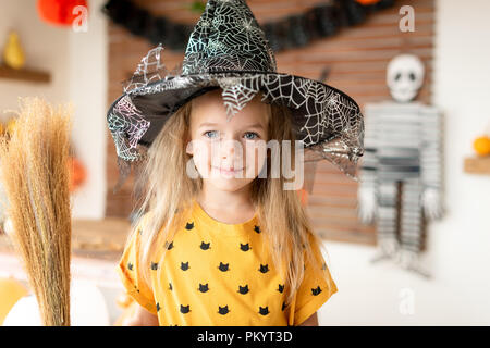 Süße kleine Mädchen in der Hexe Kostüm mit Hut hält einen Besen und Stellung im Halloween eingerichtetes Wohnzimmer, Blick auf Kamera und lächelnd. Hallowee Stockfoto