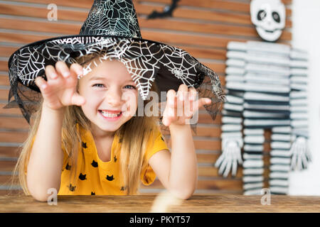 Süße kleine Mädchen mit Hexenhut hinter einem Tisch in Halloween Thema eingerichteten Wohnzimmer sitzen, an der Kamera schaut und beängstigend. Halloween Stockfoto