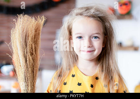 Süße kleine Mädchen mit unordentlichen Haar, verkleidet als eine Hexe und einen Besen in Halloween eingerichtetes Wohnzimmer steht, in die Kamera lächeln. Stockfoto