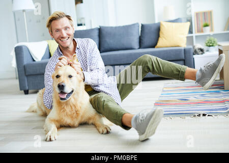 Lachende junge Mann sitzen auf dem Boden des Wohnzimmers und Genießen spielen mit flauschigen pet Stockfoto