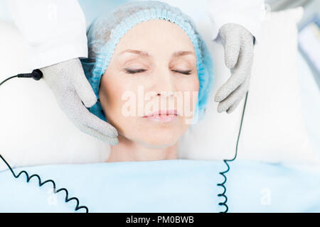 Reifen Client mit geschlossenen Augen in Spa Verfahren für die Gesichtshaut während Kosmetikerin halten Hände in thermogloves durch ihr Gesicht Stockfoto