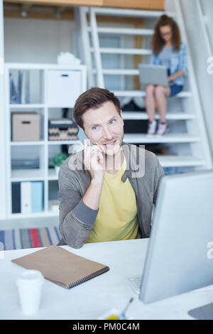 Junge erfolgreiche Designer Schreibtisch saß und mit jemand sprechen auf dem Smartphone mit Kollege networking hinter Stockfoto