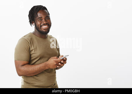 Positive charmante Dunkelhäutige männliche Modell mit Bart in militärischer Kleidung, stehend die Hälfte - gedreht mit Smartphone und breit grinsend an Kamera und genießen Sie das Surfen im Netz mit neuen Gadget Stockfoto