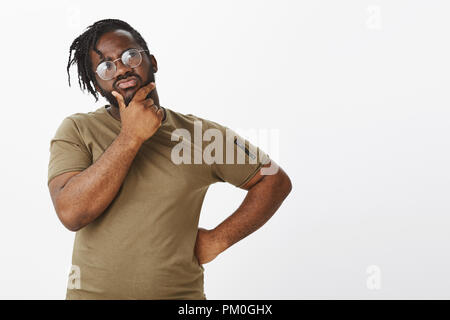 Hmm, was sollte Mann tun. Portrait von unruhigen konzentriert stattlicher Mann mit dunkler Haut in T-Shirt und Brille, die Hand auf die Hüfte und Kinn reiben während nachdenklich an der oberen rechten Ecke auf der Suche Stockfoto