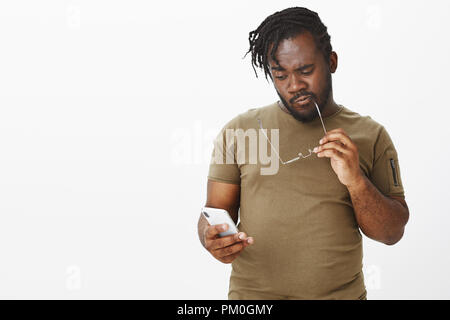 Studio shot von nachdenklich Smart afrikanischen Mann in casual Outfit, beißen Rim der Gläser halten Sie das Smartphone und den Blick auf den Bildschirm, Denken, die Entscheidung bzw. Wahl während über graue Wand stehend Stockfoto