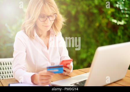 Hohe Aufnahme von attraktiven Frau mittleren Alters Einkaufen mit Kreditkarte und mit Laptop während der Sitzung in den Garten zu Hause. Stockfoto