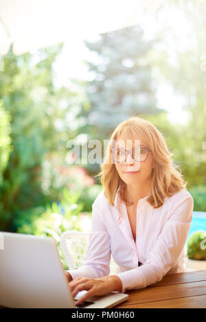 Portrait von lächelnden Frau mittleren Alters mit Ihrem Laptop im Garten, zu Hause zu sitzen. Stockfoto