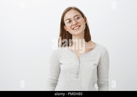 Taille-up Shot von smart Kreative junge Frau mit kurzen cute Haarschnitt Neigekopf freudig lächelnd weitgehend Brillen und casual weiße Bluse glücklich über grauer Hintergrund posiert Stockfoto