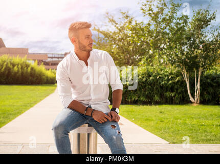 Attraktive gutaussehenden jungen Mann im City Park, Sitzen, in einem Sommertag, in Jeans und weißem T-Shirt Stockfoto