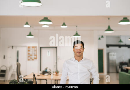 Erfolgreiche junge asiatische Geschäftsmann selbstbewusst lächelnd, während allein durch Schreibtische in einem großen modernen Büro Stockfoto