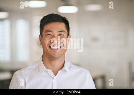 Gut gekleidete junge asiatische Geschäftsmann selbstbewusst lächelnd, während allein in einem hellen modernen Büro Stockfoto