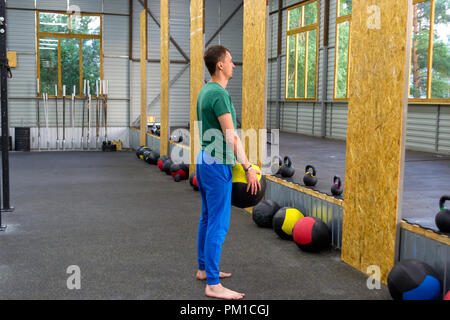 Kerl in ein grünes T-Shirt und blauen Hosen Züge in der Turnhalle, wirft eine Medizin Ball gegen die Wand, auf dem Hintergrund der Zeilen der Gewichte mit Multi-colo Stockfoto