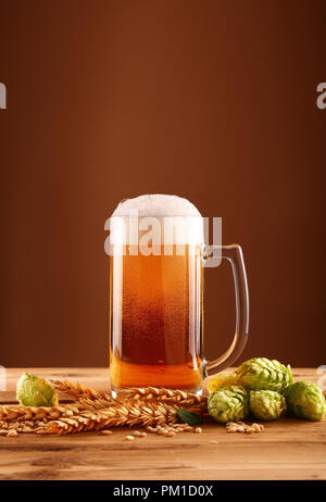 Schließen Sie ein Glas Becher Bier mit Schaum und Blasen, grün Hopfen und Gerste Korn und Spikes auf hölzernen Tisch über dunkelbraunen Hintergrund mit Kopie spa Stockfoto