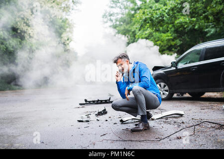 Reifer Mann einen Anruf nach einem Autounfall, Rauch im Hintergrund. Stockfoto