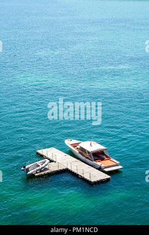 Ein Luxus Speedboot günstig zu einem isolierten, T-Form, Holz- Dock im grünen Wasser mit einem Beiboot auf dem Dock. Stockfoto